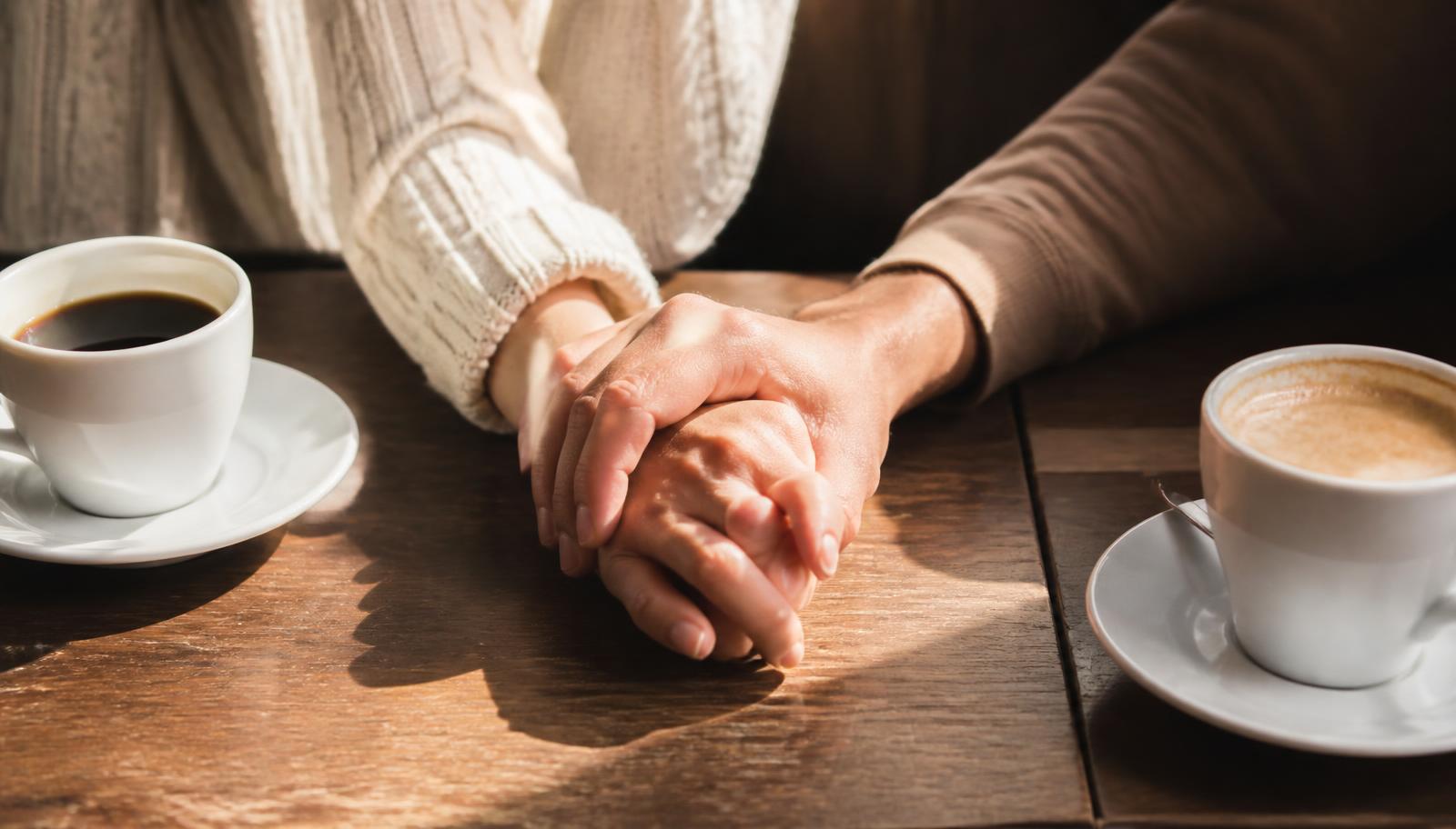 Two people's hands resting together on a sunlit wooden table beside coffee cups