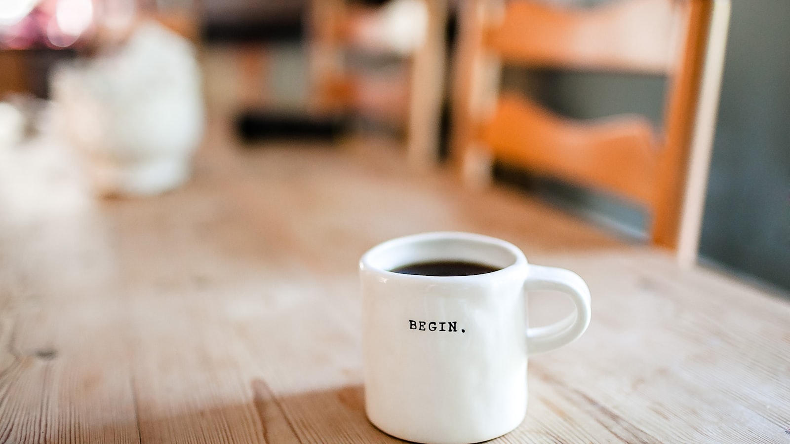 A white mug of coffee reading “Begin.” on a sunlit wooden table